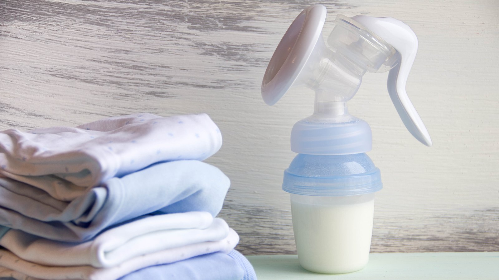 Extractor de leche manual con el biberón lleno de leche materna, junto a una pila de ropa de bebé doblada