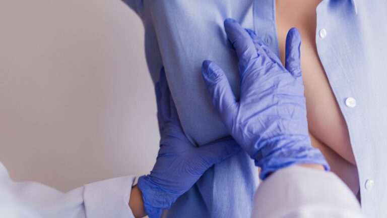 Una médica con guantes explora el pecho de una paciente que tiene la camisa abierta