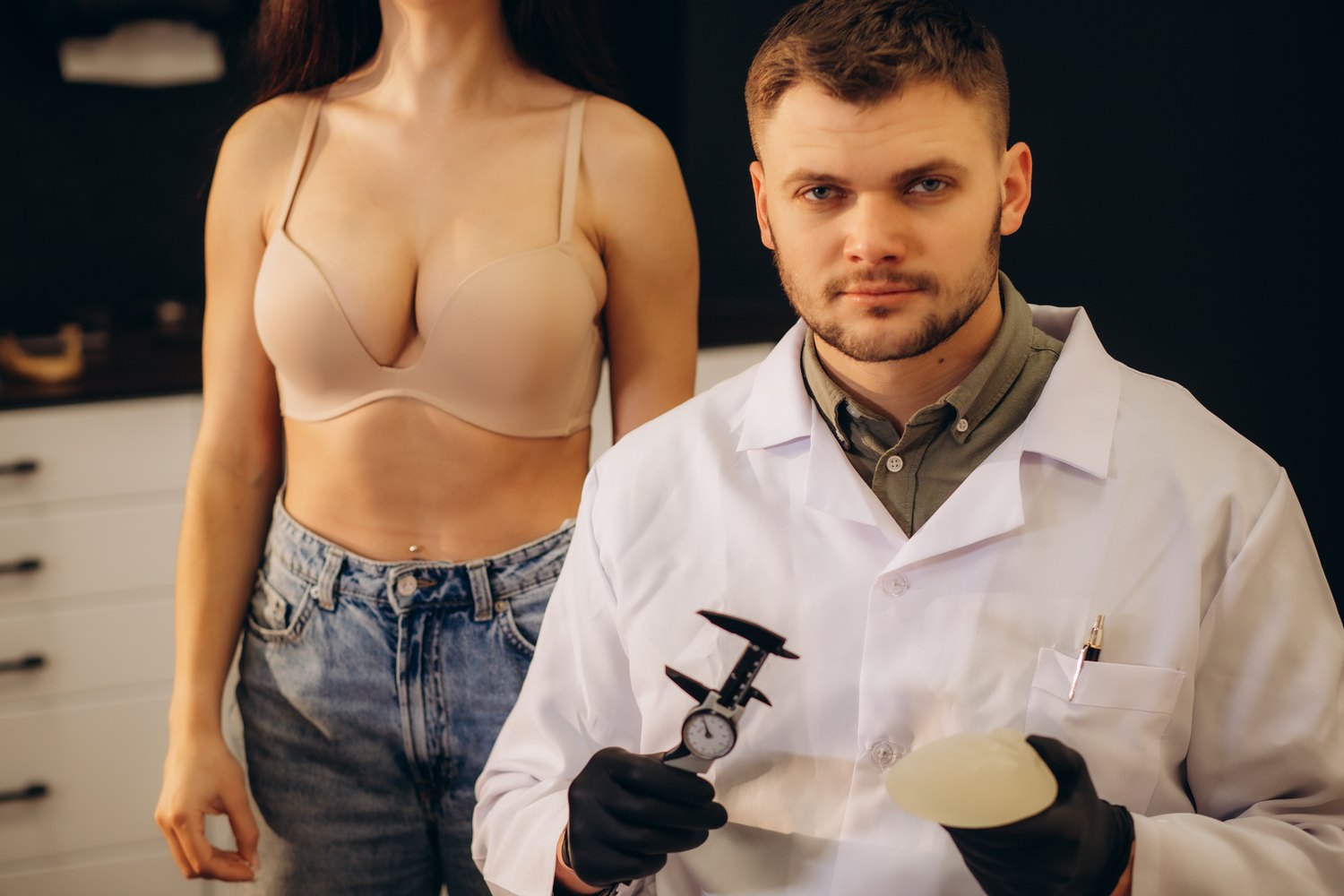 Médico con un implante de pecho y un calibre de medir, junto a una paciente en sujetador
